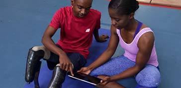 High angle front view of an African Amercian man with two prosthetic legs wearing sports clothes at a sports centre gym sitting on the floor and looking at a tablet computer with an African American female physiotherapist and smiling