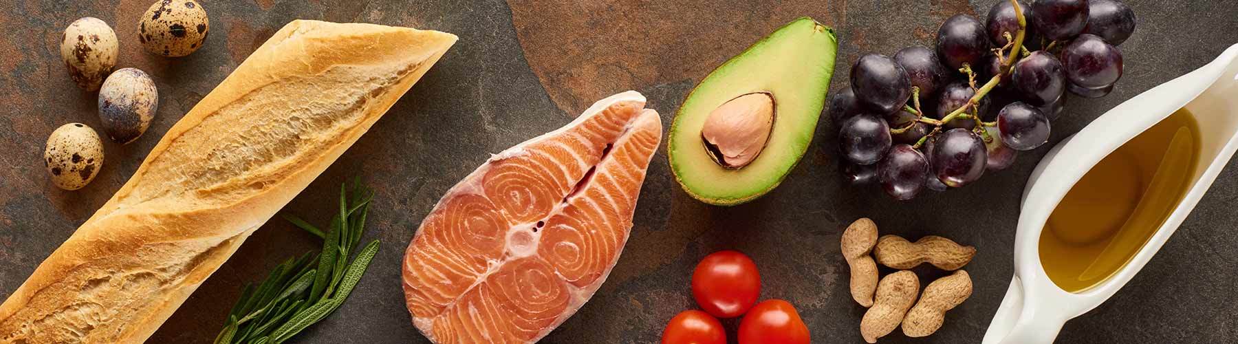 Top view of raw fish near vegetables, fruits, eggs, peanuts, baguette, herb twigs and olive oil on marble surface with copy space
