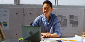 Front view of Asian male architect using laptop on desk in a modern office. He is sitting at desk