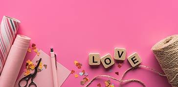 Top view of valentines decoration, scissors, wrapping paper, twine and love lettering on wooden cubes on pink background